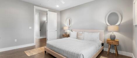 A bedroom with a bed, two bedside tables with lamps, a ceiling fan, and a door leading to another room.