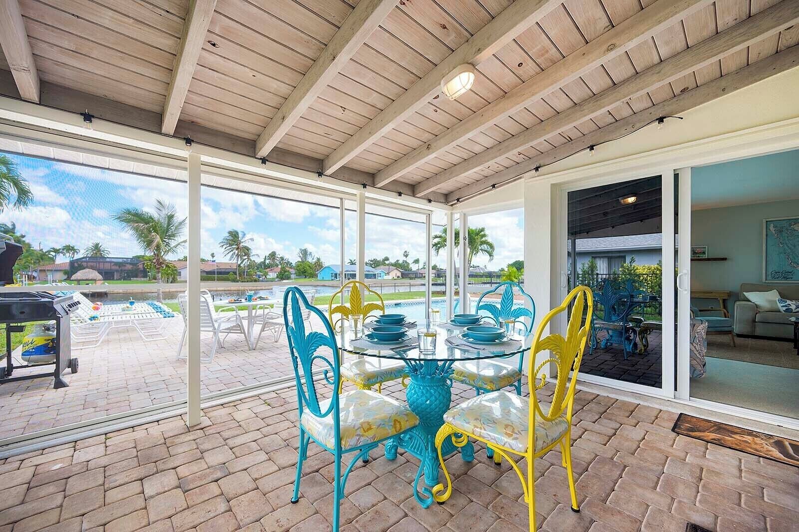 A covered patio with a glass table and chairs, a grill, and a view of a swimming pool and palm trees.