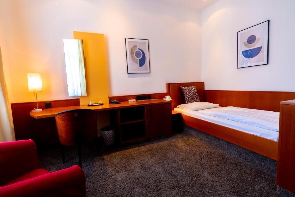 Habitación de hotel con cama, escritorio, silla, lámpara y dos obras de arte abstractas en la pared.