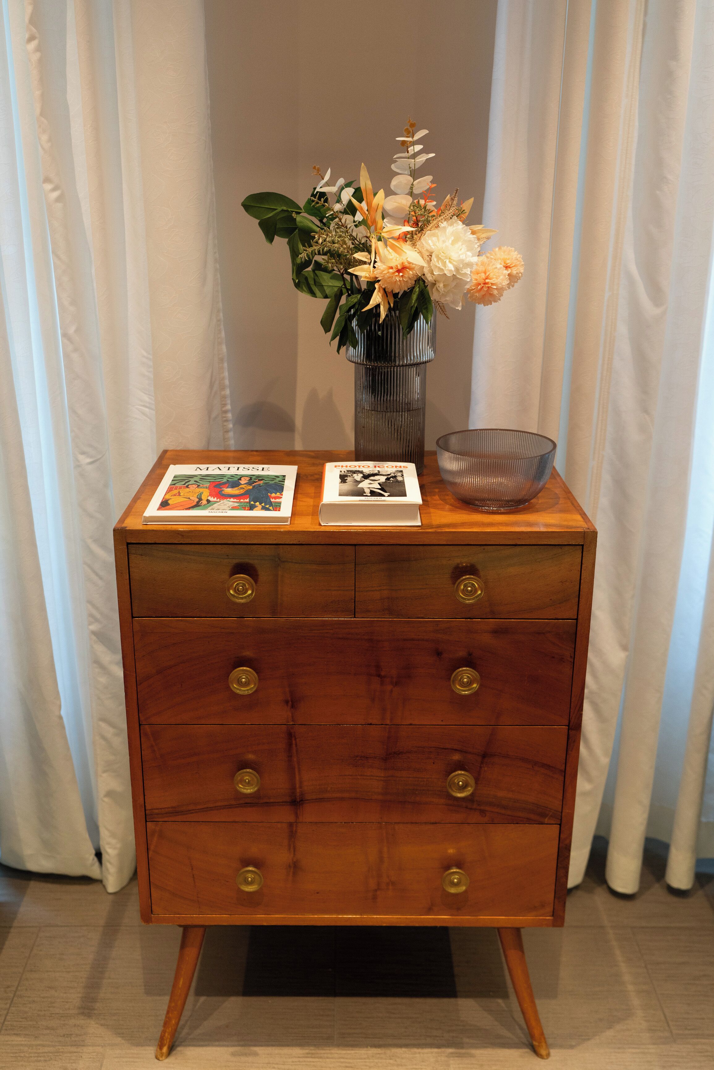 A wooden dresser with a vase of flowers, two books, and a glass bowl.
