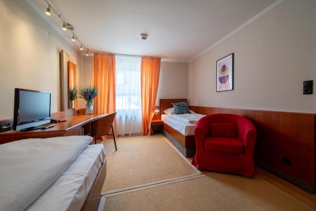 Ett hotellrum med en säng, ett skrivbord med en dator, en röd stol, ett litet bord med en växt och en tavla på väggen.