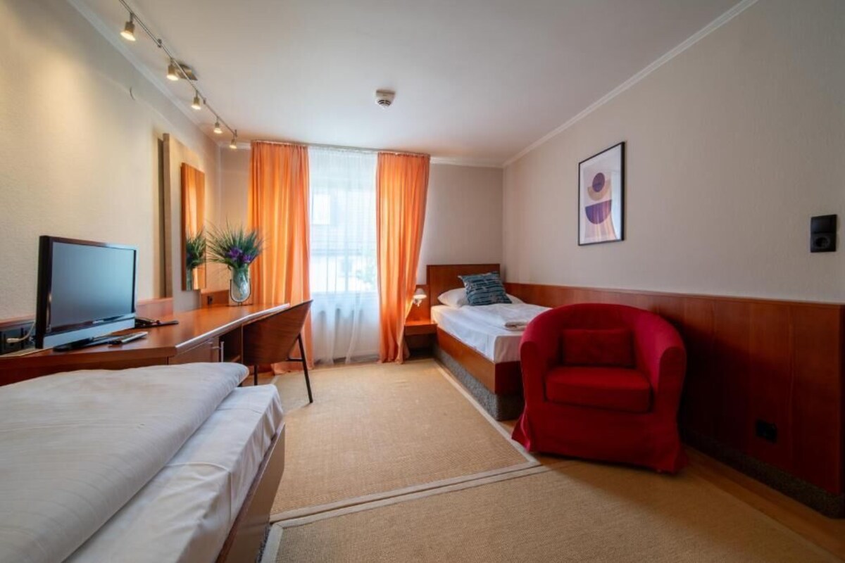 Una habitación de hotel con cama, escritorio con computadora, una silla roja, una pequeña mesa con planta y un cuadro en la pared.