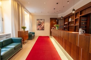 Una hall d'albergo con tappeto rosso, una reception, scaffali in legno con oggetti esposti, un divano verde e un quadro appeso al muro.