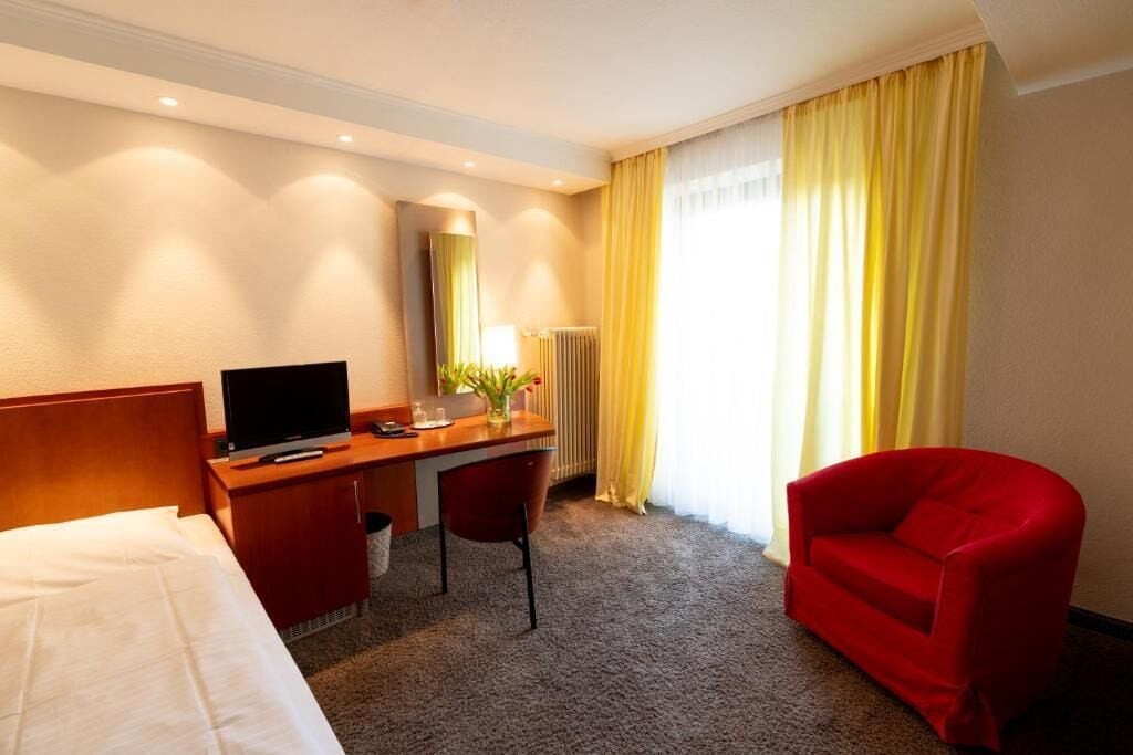 A hotel room with a bed, a desk with a chair, a television, a radiator, and a window with curtains.
