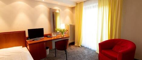 A hotel room with a bed, a desk with a chair, a television, a radiator, and a window with curtains.