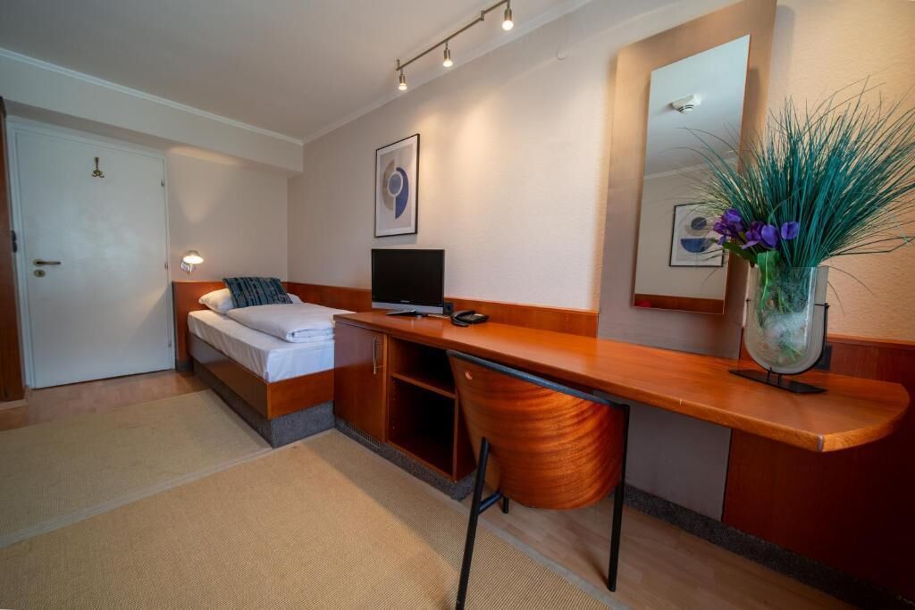 A hotel room with a bed, a desk, a chair, a television, a mirror, and a vase with flowers.