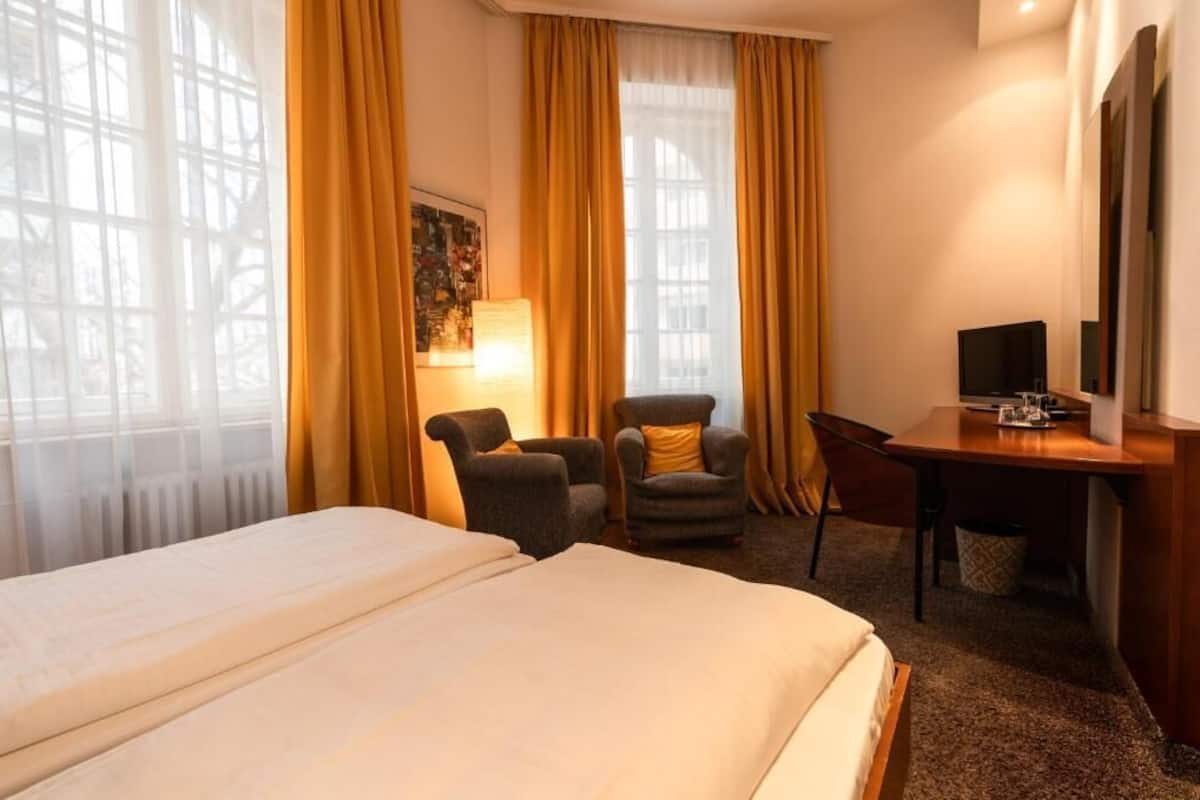 Una camera d'albergo con un letto, due poltrone, una scrivania, un televisore e un'ampia finestra con tende.