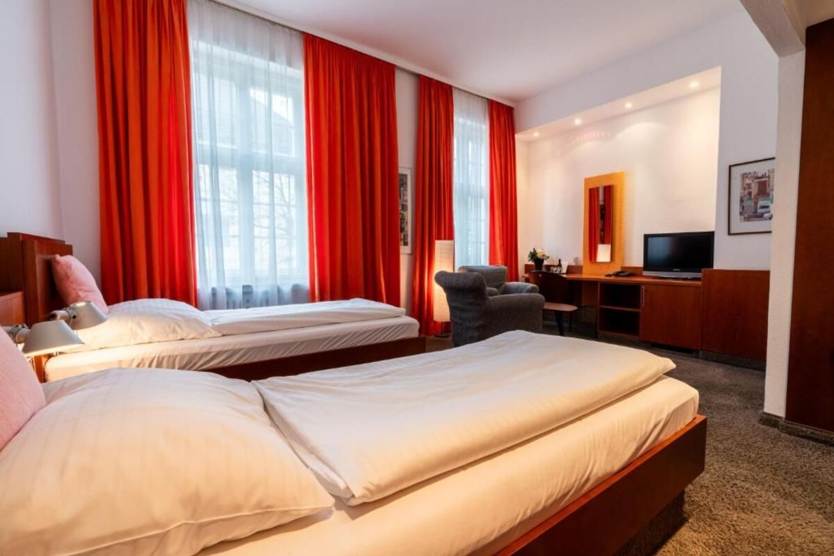 Habitación de hotel con dos camas, un escritorio, una silla, un televisor y un ventanal grande con cortinas rojas.