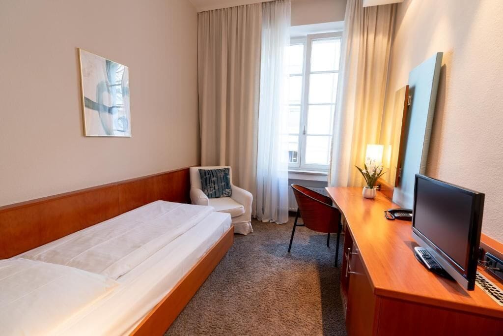 Ein Hotelzimmer mit einem Bett, einem Schreibtisch mit Computer, einem Sessel, einem Fernseher und einem Fenster mit Vorhängen.