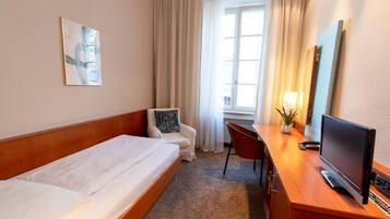Ein Hotelzimmer mit einem Bett, einem Schreibtisch mit Computer, einem Sessel, einem Fernseher und einem Fenster mit Vorhängen.
