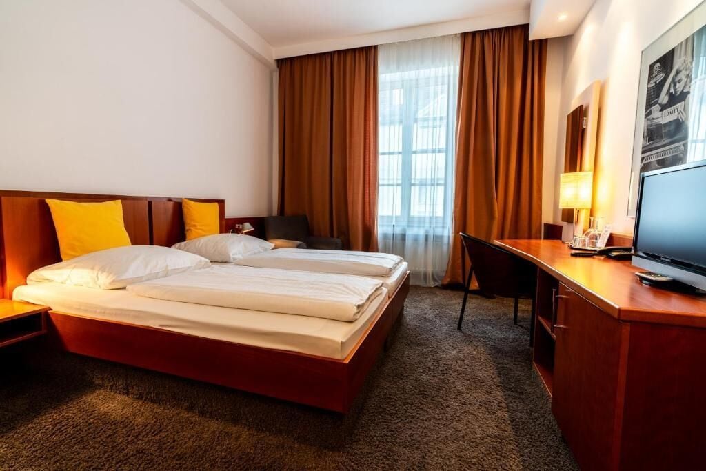 Una camera d'albergo con un letto matrimoniale, una scrivania con televisione, una sedia e una finestra con tende.