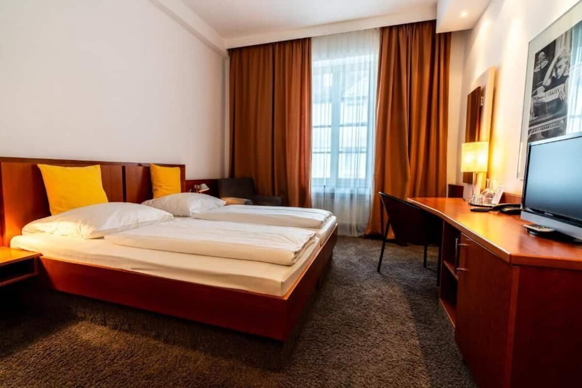 Una camera d'albergo con un letto matrimoniale, una scrivania con televisione, una sedia e una finestra con tende.