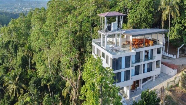 A modern multi-story building with a glass facade, surrounded by dense forest and hills in the background. - Iris - Kandy (Kandy)