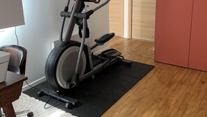 A home gym with an elliptical machine, a desk with a laptop, a chair, a rug, and a wardrobe.