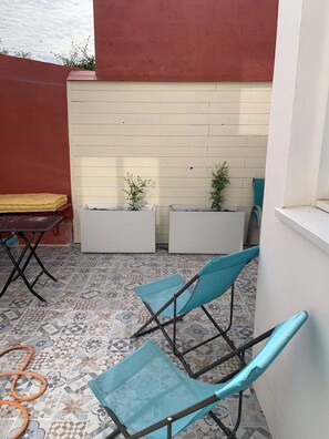A patio with a patterned tile floor, two blue folding chairs, a yellow cushioned bench, and two white planters with small plants. - MAISON DE KERNERS (Arzon)