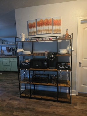 A kitchen with a wooden shelf unit holding kitchenware, a coffee bar, and a microwave.