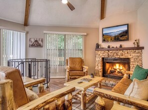A living room with a stone fireplace, wooden furniture, a television, and a view of a landscape through the window. - Octagon Chalet: Hot Tub, Pool Table & Stunning Mountain Views - Sleeps 6 (Gatlinburg)