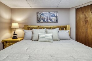 Ein Schlafzimmer mit Holz Kopfteil, einem Bett mit geripptem weißen Bettbezug, zwei Kissen, einem Nachttisch mit Lampe und einer Hirschfigur sowie einem an der Wand hängenden Bärenbild.