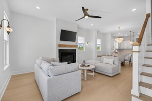 Ein geräumiges Wohnzimmer mit grauen Sofas, einem Kamin, einem Flachbildfernseher und einer Küche im Hintergrund.