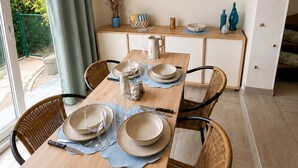 A dining area with a wooden table set for four, chairs, a small side table with decorative items, and a view of the outdoors through a glass door. - Family home in secure residence with swimming pools close to beaches (Béziers)