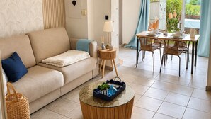 A living room with a sofa, a round coffee table, a dining area with a table and chairs, and a view of greenery outside. - Family home in secure residence with swimming pools close to beaches (Béziers)