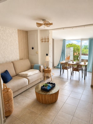 A living room with a sofa, a round coffee table, a dining area with a table and chairs, and a view of greenery outside. - Family home in secure residence with swimming pools close to beaches (Béziers)