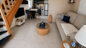 A modern living room with a wooden staircase, a sofa, a coffee table, and a kitchen area in the background. - Family home in secure residence with swimming pools close to beaches (Béziers)