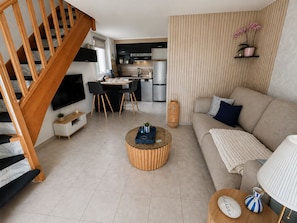 A modern living room with a wooden staircase, a sofa, a coffee table, and a kitchen area in the background. - Family home in secure residence with swimming pools close to beaches (Béziers)