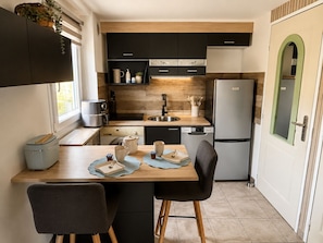 A compact kitchen with modern appliances, a dining area with two chairs, and a view of the outdoors through a window. - Family home in secure residence with swimming pools close to beaches (Béziers)
