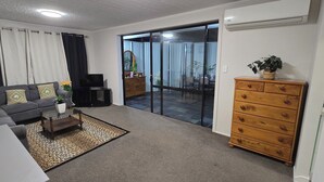 A living room with a sofa, a coffee table, a TV, a dresser, and a sliding glass door leading to a balcony. - Modern 2BR Home, Sleeps 5,  Parking,  Near Eastgate (Christchurch)