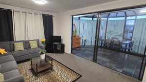 A living room with a sofa, coffee table, TV, and a sliding glass door leading to an outdoor area with a table and chairs. - Modern 2BR Home, Sleeps 5,  Parking,  Near Eastgate (Christchurch)