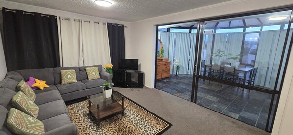 Ein Wohnzimmer mit Sofa, Couchtisch, Fernseher und einer Schiebetür zu einem Außenbereich mit Tisch und Stühlen.