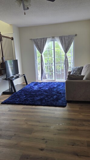 Ein Wohnzimmer mit blauem Teppich, einem Fernseher auf einem Schrank, einer Couch und einem Fenster mit Vorhängen.