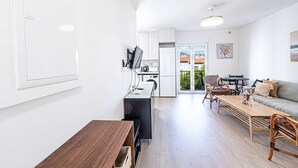 Cozinha moderna com armários brancos, área de jantar com mesa e cadeiras de madeira, e sala de estar com sofá e televisão.