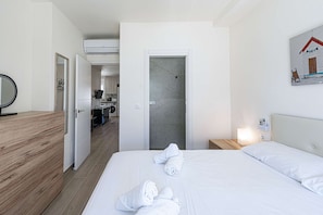 Ein Hotelzimmer mit Holzboden, einem Bett mit weißer Bettwäsche, einem Badezimmer mit Dusche und einer sichtbaren Küchenzeile durch eine offene Tür.