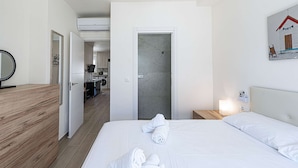 A hotel room with a wooden floor, a bed with white linens, a bathroom with a shower, and a kitchen area visible through an open door. - Hacienda 4A by Casasol (Nerja)