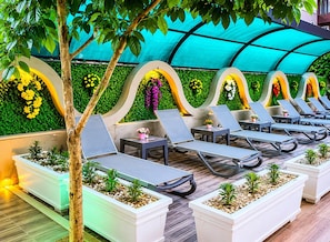 A sunlit outdoor lounge area with lounge chairs and planters, under a curved roof structure. - LEON OTEL (Manavgat)