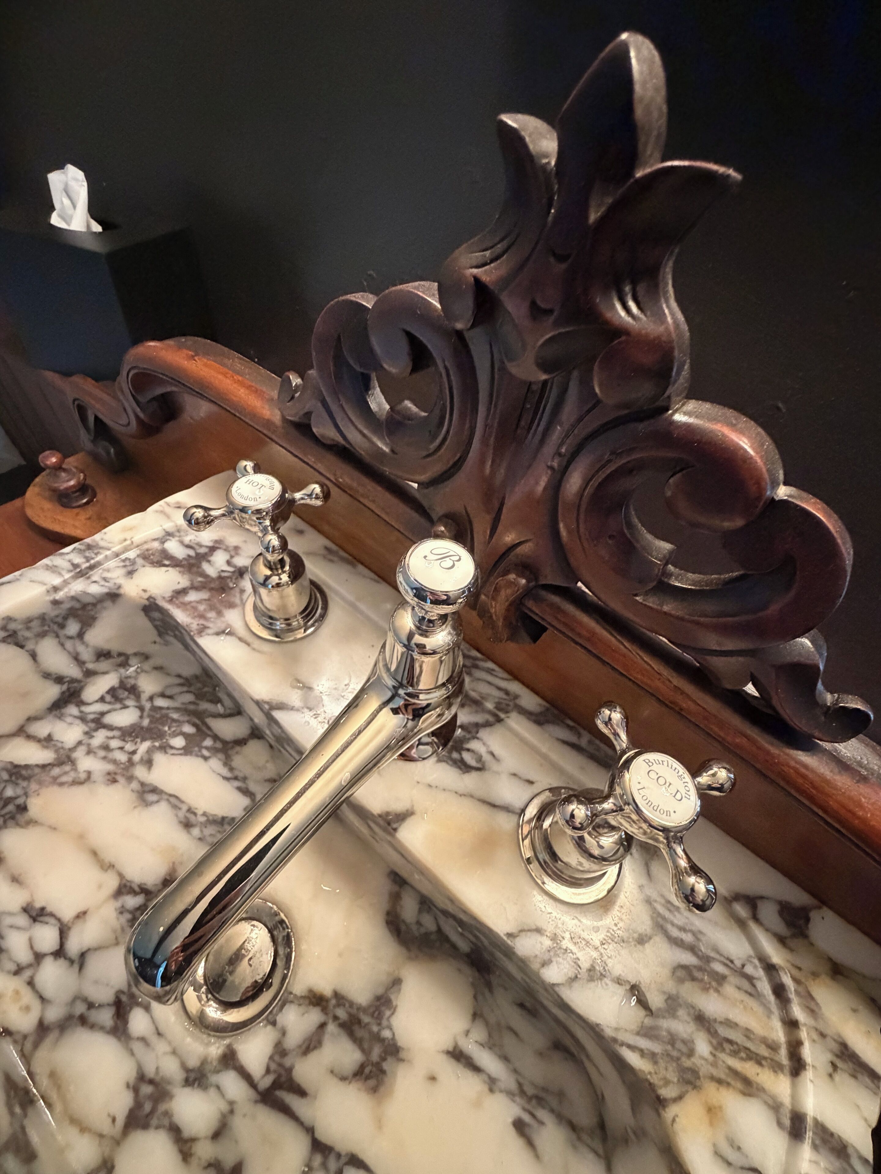 Un baño con un lavabo de mármol, dos grifos y un espejo de madera con adornos decorativos.