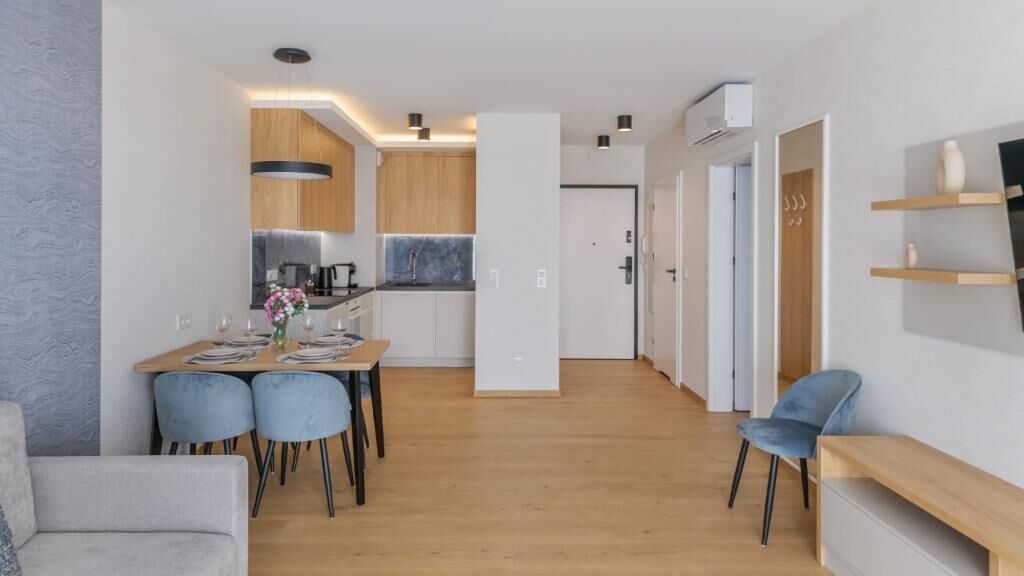 A modern kitchen with wooden cabinets, a dining area with blue chairs, and a living area with a sofa and TV.
