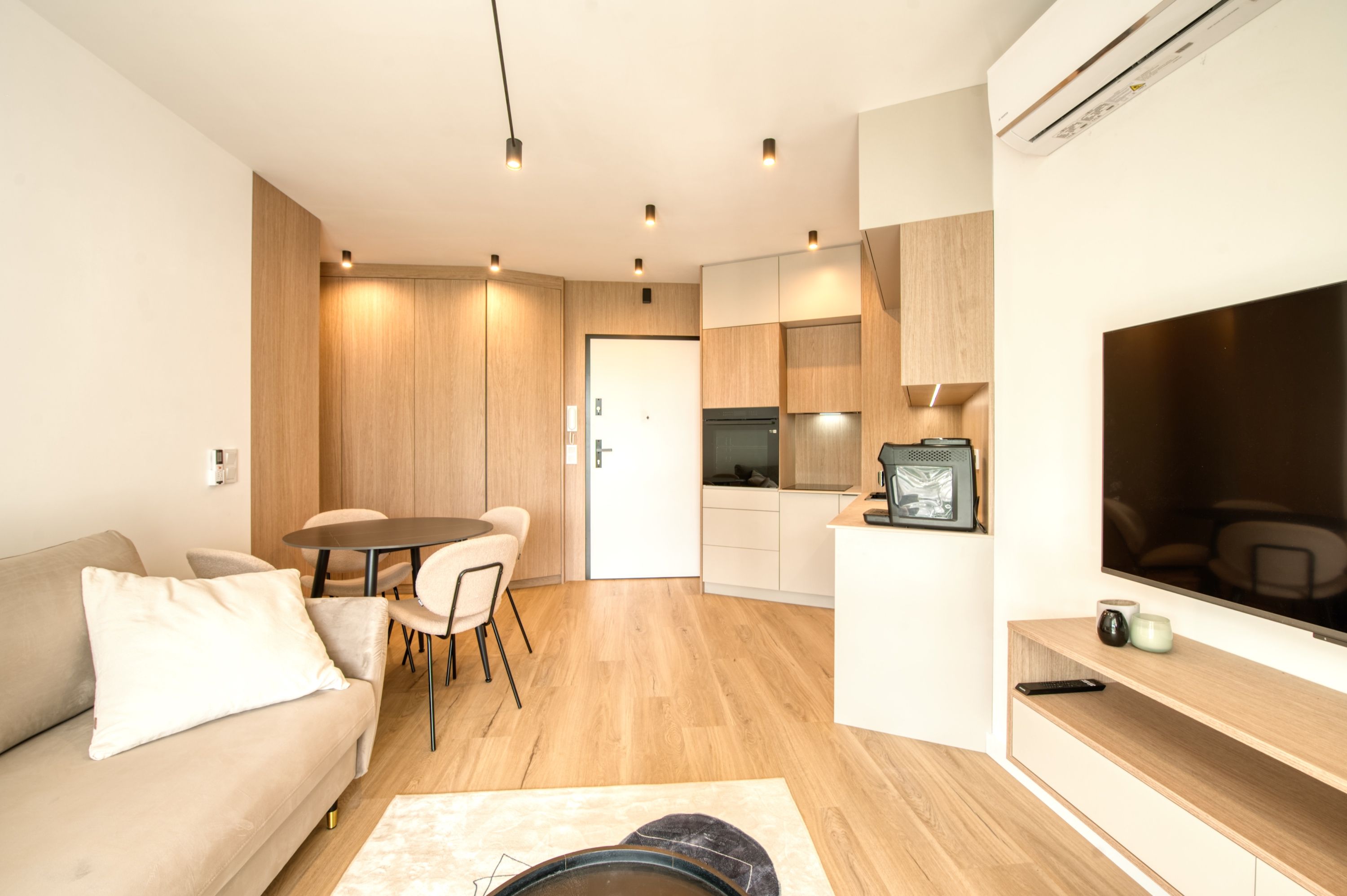 A modern living room with a sofa, a dining table, and chairs. There is a kitchen area with a microwave and a TV.