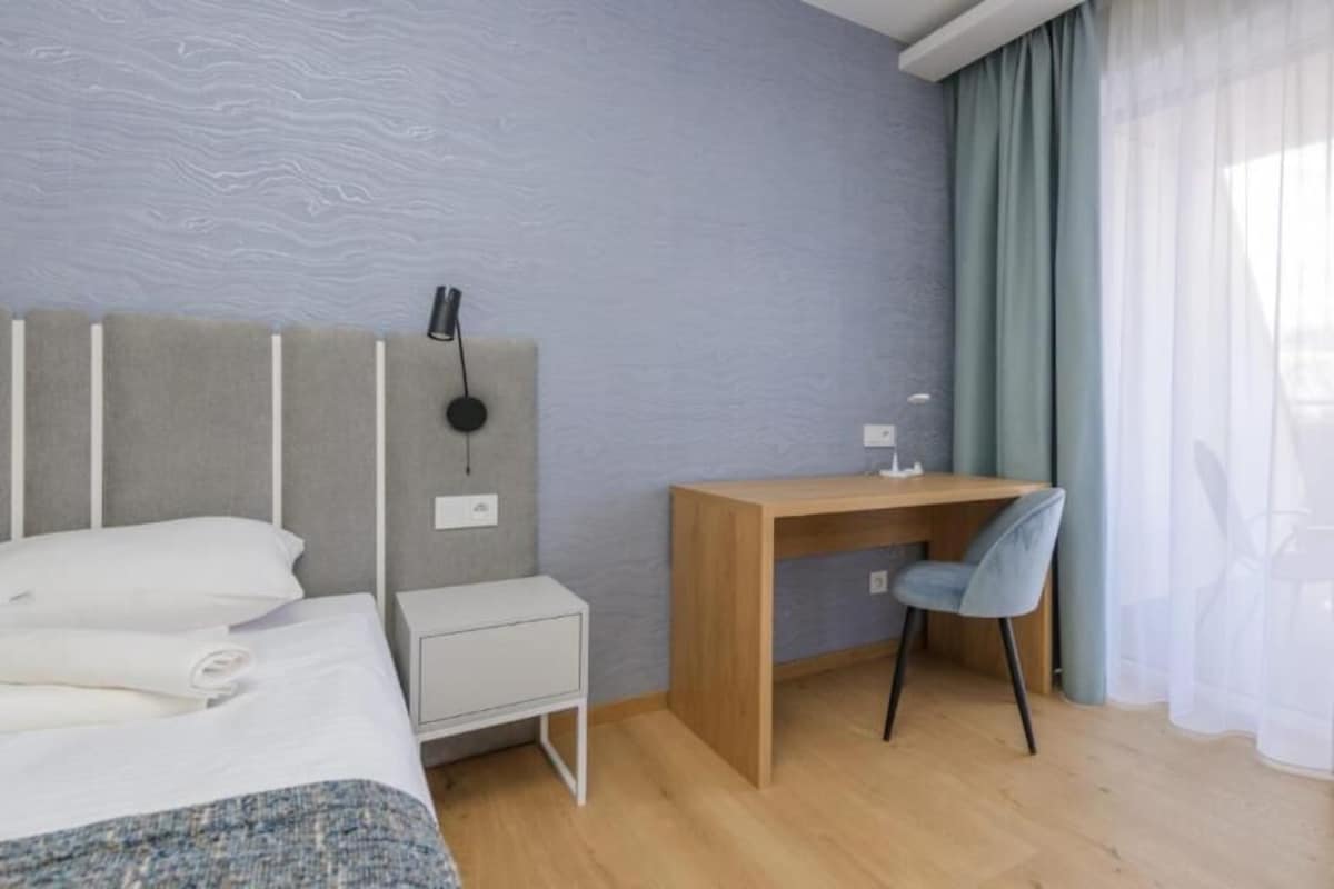Una camera d'albergo moderna con un letto, una scrivania, una sedia, un comodino e una lampada a parete.