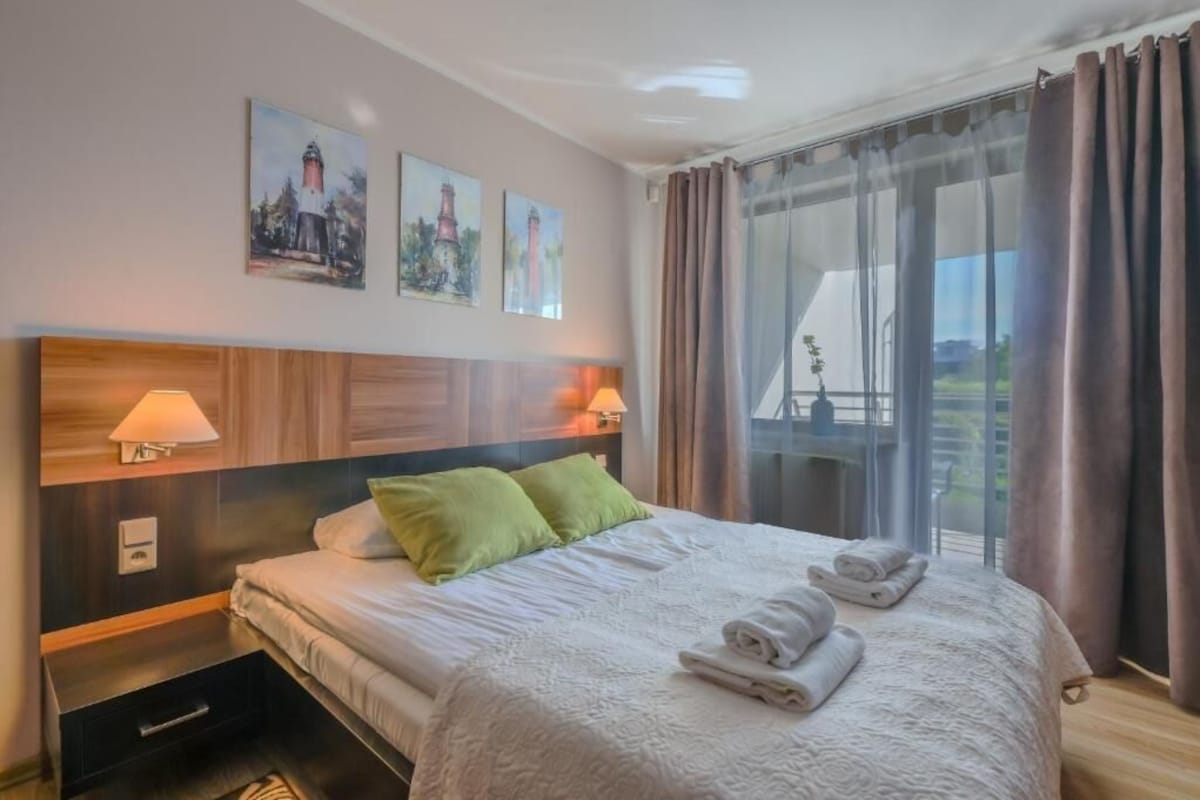 Una camera d'albergo con un letto, due cuscini, un comodino, una lampada, una finestra con tende e un'opera d'arte appesa al muro.