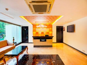 Un área de recepción de hotel con diseño moderno, que incluye un mostrador de recepción, un sofá, una mesa de centro y un televisor montado en la pared.