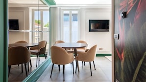 Een moderne kamer met een ronde tafel en stoelen, een flatscreen televisie aan de muur en een groot raam met gordijnen.