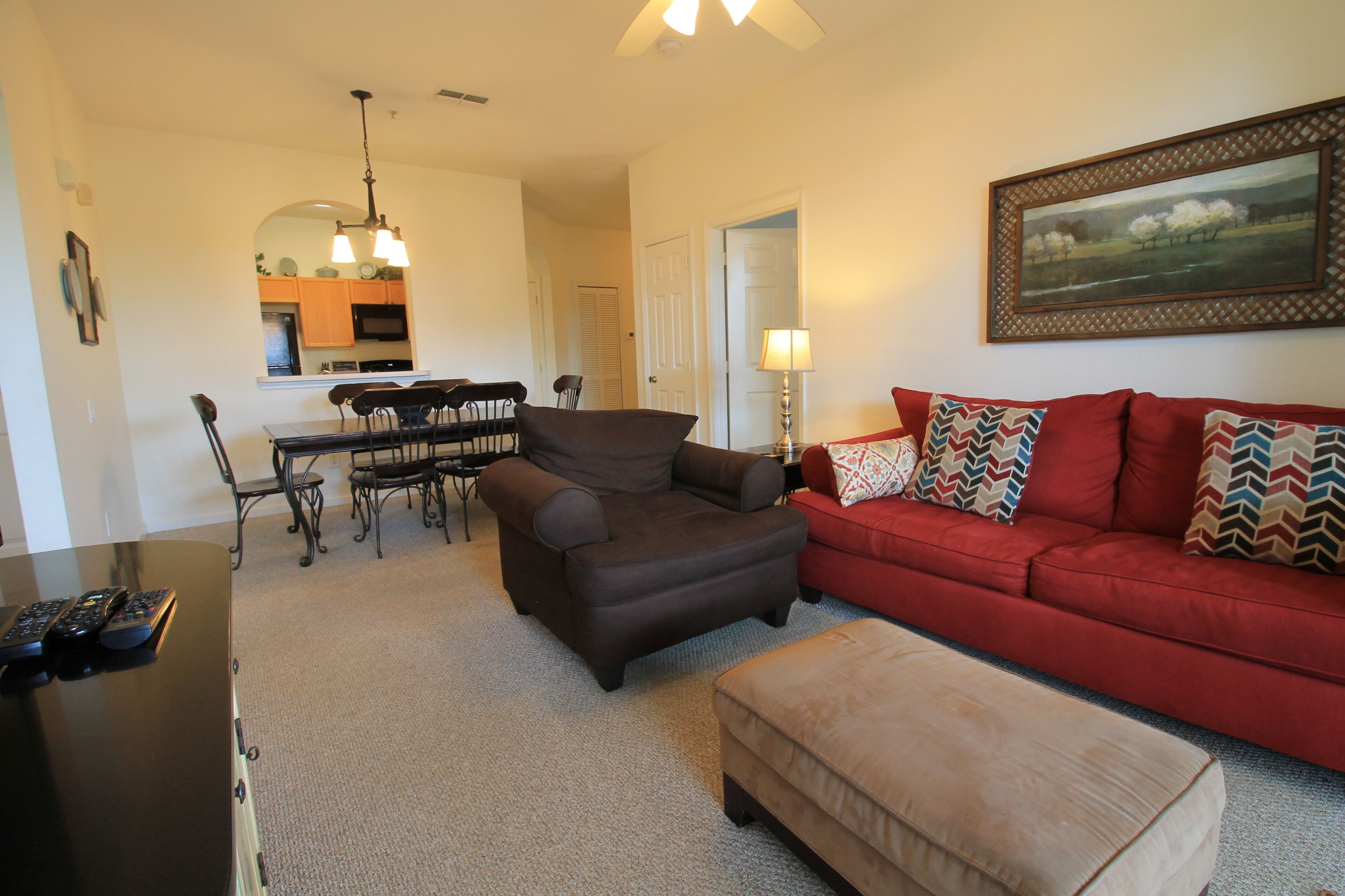 A living room with a red sofa, a brown armchair, a beige ottoman, a dining area with a table and chairs, and a kitchen in the background.