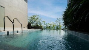 Una piscina exterior con barandillas metálicas, borde de hormigón y vistas a árboles y un edificio al fondo.