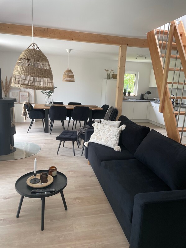 A modern living room with a dark sofa, a dining table with chairs, a hanging wicker light fixture, and a staircase.