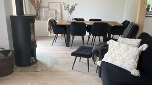 A modern living room with a black sofa, a round coffee table with remote controls, a black fireplace, a dining area with a wooden table and chairs, and a hanging wicker lamp. - Holiday Home 'Strand Haus' with Private Terrace, Private Garden and Wi-Fi (Elsfleth)