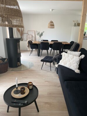 A modern living room with a black sofa, a round coffee table with remote controls, a black fireplace, a dining area with a wooden table and chairs, and a hanging wicker lamp.