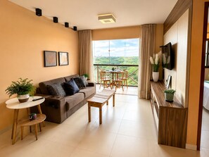 A modern living room with a sofa, coffee table, and dining table. There is a TV mounted on the wall, and a balcony with a view.